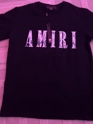 Svart AMIRI t-shirt med tryck - Svart t-shirt från AMIRI med stort vitt och blått logotryck på bröstet. Klassisk rund halsringning och korta ärmar. Tillverkad i mjuk bomull som känns skön mot huden. Perfekt för dig som gillar streetwear och vill sticka ut med en snygg logga.
