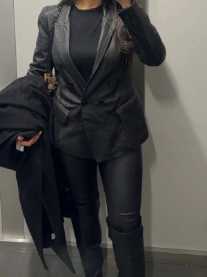 Svart skinnkavaj med dubbelknäppning - Snygg svart kavaj i skinnimitation med dubbelknäppning och klassisk krage. Kavajen har två fickor framtill och en figurnära passform som ger en cool och stilren look. Perfekt för dig som vill sticka ut med en edgy vibe.