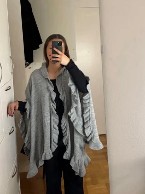 Grå poncho med volangdetaljer - Säljer en stilren grå poncho med volangdetaljer. Perfekt för lager-på-lager och ger en elegant touch till din outfit. Den är onesize och passar de flesta. Super fint skick, använd endast 2 gånger! 