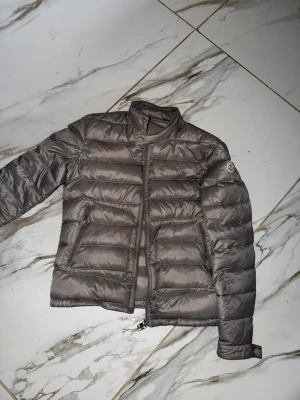 Grå dunjacka från Moncler - Snygg grå dunjacka från Moncler med quiltad design och ståkrage. Jackan har dragkedja framtill som har förstörts lite men funkar ifall du drar upp den lungt. två sidofickor med dragkedja och en diskret Moncler-logga på ärmen. Tillverkad i glansigt material och fylld med dun för extra värme. Passar xs-s