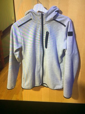 Ljusgrå hoodie  med huva - Ljusgrå hoodie med hel dragkedja framtill, svart bröstficka med dragkedja och huva. Svarta detaljer på axlar och ärm, samt en svart patch på vänster ärm. Hoodie har lång ärm och sportig look.
