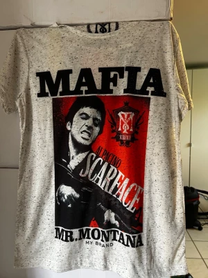 Vit t-shirt Scarface Mr. Montana - Cool vit t-shirt med svart och rött Scarface-tryck framtill och emblem på ryggen. Stora bokstäver 'MAFIA' och 'SCARFACE' på framsidan, samt detaljerad grafik. T-shirten är melerad och har rund hals. Perfekt för dig som gillar streetwear och filmreferenser.