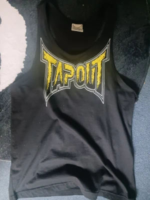 Svart Tapout linne med tryck - Snyggt svart linne från Tapout med stort gult och vitt logotryck framtill och mindre logga på ryggen. Perfekt för gymmet eller till vardagslooken. Tillverkat i mjukt och bekvämt material som andas. Klassisk ärmlös modell med rundad hals.