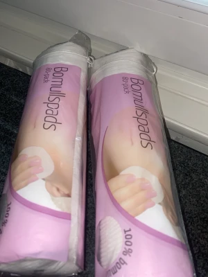Bomullspads 80-pack - Två förpackningar bomullspads, 80 stycken i varje, perfekta för sminkborttagning eller ansiktsrengöring. Padsen är vita, runda och tillverkade av 100% bomull. Förpackningen är rosa och vit med tydlig text.