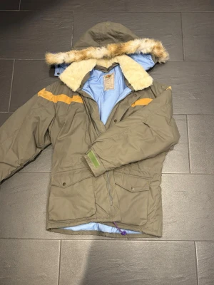 Olivgrön vintage Fjällräven parkas med fuskpäls och huva - Olivgrön parkas med ljusblått foder, avtagbar huva och fuskpäls runt kragen. Jackan har gula detaljer på axlarna, stora fickor framtill och dragkedja med tryckknappar. Perfekt för kalla dagar och riktigt snygg streetstil. Helt oanvänd Fjällräven jacka, nypris 7000kr+, storlek L. Säljer den för 3000kr. 