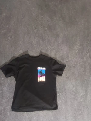 Svart Miami t-shirt med tryck - Cool svart t-shirt med färgglatt Miami-tryck både fram och bak. Motivet visar palmer mot en solnedgång i blått, rosa och orange. Perfekt för dig som gillar streetstyle och grafiska prints.