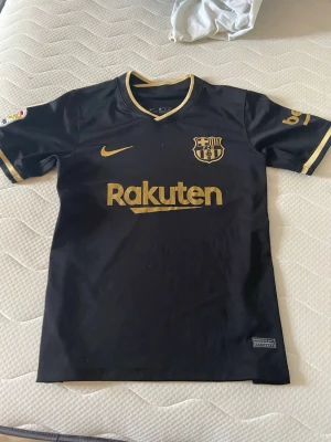 Svart FC Barcelona matchtröja Nike - Snygg svart FC Barcelona matchtröja från Nike med guldfärgade detaljer och Rakuten-logga på bröstet. Tröjan har klubbmärke och Nike-logga i guld, samt guldfärgade ränder på ärmarna. Perfekt för dig som älskar fotboll och vill representera Barca.