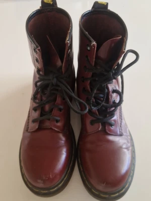 Vinröda Dr. Martens kängor i läder - Säljer ett par klassiska vinröda Dr. Martens kängor i äkta läder. De har svarta snören, högt skaft och den ikoniska gula sömmen runt sulan. Perfekta för dig som vill ha en tidlös och edgy stil.