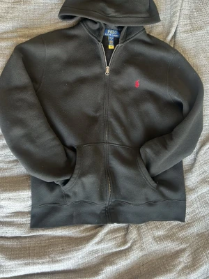 Svart hoodie från Polo Ralph Lauren - Svart hoodie från Polo Ralph Lauren med dragkedja framtill och klassisk huva. Har en liten röd broderad logga på bröstet och stora fickor framtill. Perfekt för en chill och stilren look. Sitter som en xs-s på man skulle jag säga 😌använd men inga skador!