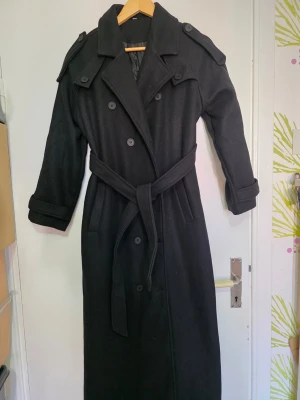 Svart lång trenchcoat med bälte - Stilren svart trenchcoat med dubbelknäppning, axelklaffar och avtagbart midjebälte. Långa ärmar med knäppning vid mudden och klassisk krage. Tillverkad i ett tjockare tyg som passar perfekt för kyligare dagar. Två sidofickor och slits baktill för extra rörelsefrihet.