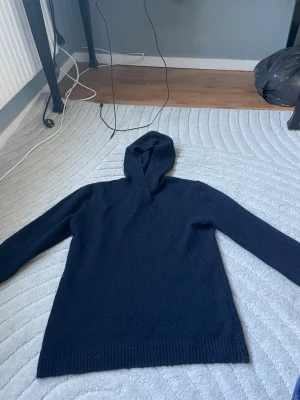 Mörkblå stickad hoodie - Säljer en mörkblå stickad hoodie med ribbade muddar och huva. Tröjan har en enkel och clean design utan tryck eller logga, perfekt för dig som gillar en stilren look. Materialet känns mjukt och passar bra till både jeans och mjukisbyxor.