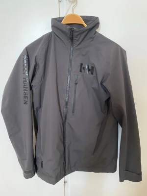Mörkgrå Helly Hansen vindjacka - Snygg mörkgrå vindjacka från Helly Hansen med hög krage, dragkedja framtill och logga på bröstet samt ärmen. Jackan har en bröstficka med dragkedja och en gul detalj på innerfickan. Tillverkad i slitstarkt material, perfekt för blåsiga dagar.