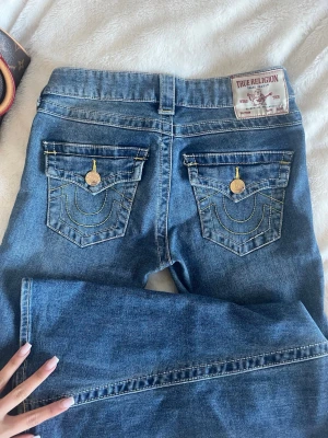 Blå True Religion jeans med fickdetaljer - Säljer ett par blå jeans från True Religion med ikoniska bakfickor, gul kontrastsöm och guldiga knappar. Klassisk femficksmodell i denim med patch på linningen. Perfekta för dig som gillar streetstyle och vill sticka ut med snygga detaljer.