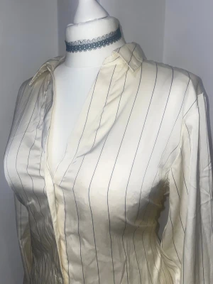 Randig vit skjorta i satin - Snygg off white/kräm vit skjorta med tunna vertikala ränder och klassisk krage. Skjortan har lång ärm och en glansig finish i satin. Perfekt att styla till jeans, kan stylas i oversized eller mera fitted med klämma eller knut i ryggen.