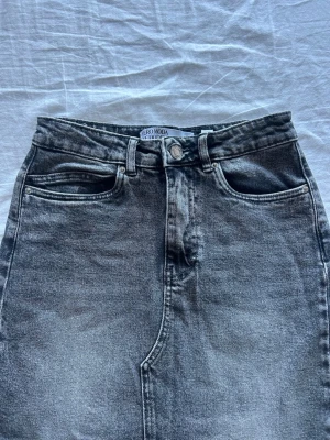 Svartgrå jeanskjol från Vero Moda - Säljer en svartgrå jeanskjol från Vero Moda med knapp och dragkedja framtill. Kjolen har en tvättad look och är lång med en slit längst ner vid benen. Storlek xs