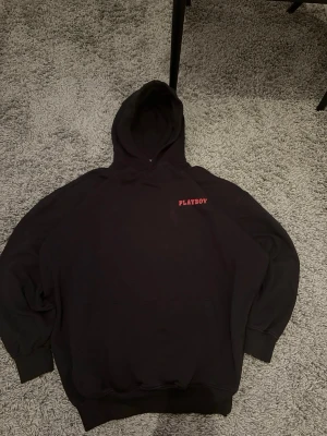Svart hoodie Playboy x Missguided - Säljer en svart hoodie från Playboy x Missguided med röd PLAYBOY-text på bröstet. Klassisk oversized passform med huva och magficka. Tillverkad i mjukt material, perfekt för en chill och trendig look.