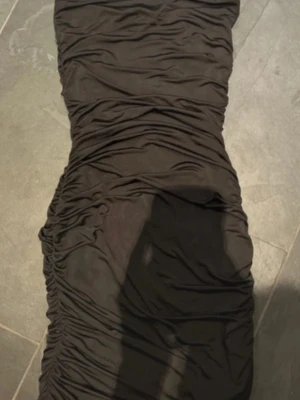 Svart omlottklänning med snörning - Säljer en svart kortklänning med smala axelband och snygg omlottdesign. Klänningen har rynkade detaljer längs hela plagget och snörning i ryggen för en justerbar passform. Perfekt för dig som gillar en trendig och figurnära look.