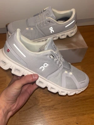 Grå On Cloud 6 sneakers storlek 43 - Snygga grå On Cloud sneakers med vita detaljer och reflekterande inslag. Ovandelen är i mesh och syntet för bra andningsförmåga och komfort. Sulan är vit och har On's unika CloudTec-design för extra dämpning. Perfekta för en sportig och modern look.