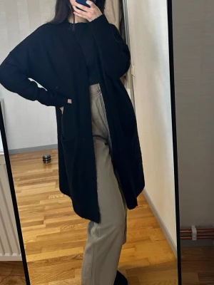 Svart lång kofta från 157 - Mysig svart lång kofta från 157 med öppen front och två fickor framtill. Koftan är stickad och har långa ärmar samt ribbade muddar vid ärmslut och nederkant. Perfekt att slänga över en t-shirt eller hoodie för en chill look.