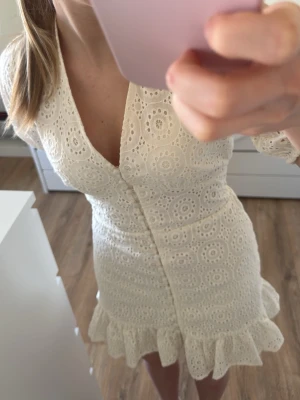 Vit spetsklänning med volang - Söt vit kortklänning med v-ringning, knappar framtill och volangkant nedtill. Klänningen har ett broderat spetsmönster och korta ärmar. Perfekt för sommarens alla tillfällen!