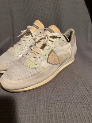  Philippe Model - Snygga beige sneakers från Philippe Model med detaljer i mocka och textil. Skorna har vita snören, en beige sula och skimrande partier vid snörningen. Klassisk låg modell med logotyp på sidan och bekväm passform.