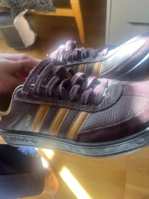 Adidas Madrid vinröda sneakers - Snygga Adidas Madrid sneakers i vinrött med klassiska guldiga ränder på sidorna. Ovandelen är i läder och mocka, med matchande vinröda skosnören och en slitstark gummisula. Perfekta för dig som gillar retrostil och streetwear.