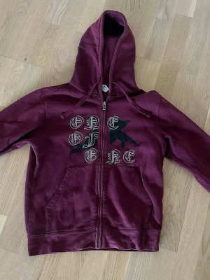 Vinröd hoodie med tryck och dragkedja - Säljer en vinröd hoodie med dragkedja och huva. På framsidan står det 'ONE OF ONE' i vit gotisk stil och ett svart motiv bakom texten. Hoodien har känguruficka och justerbara snören i huvan. Materialet känns mjukt och bekvämt. Tröjan säljs inte längre och är i limiterat antal.