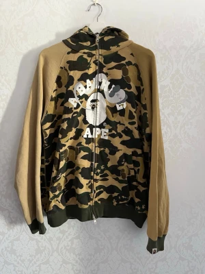 A Bathing Ape camo zip hoodie - Säljer en camo-hoodie från A Bathing Ape med dragkedja framtill och stor vit logga på bröstet. Hoodien har gröna och bruna camouflagepaneler, enfärgade beige ärmar och muddar. Klassisk huva och ribbade avslut. Perfekt för dig som gillar streetwear och vill sticka ut.