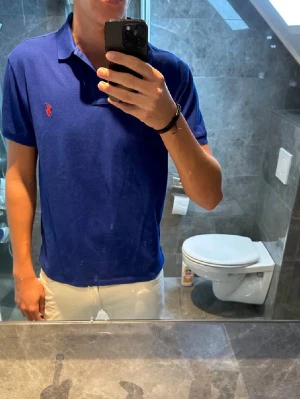 Blå piké från Ralph Lauren - Klassisk blå pikétröja från Ralph Lauren med den ikoniska röda loggan broderad på bröstet. Tröjan har krage, korta ärmar och knappar vid halsen. Perfekt till jeans eller shorts för en clean och stilren look.