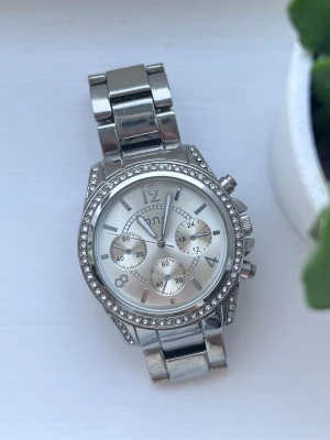 Silvrig klocka med strassdetaljer - Snygg silverfärgad klocka från O.N.E med strass runt urtavlan. Superfin accessoar som funkar till allt! Kan behöva ett nytt batteri.