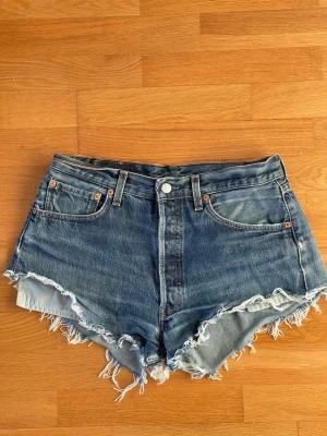 Levi's 501 blå jeansshorts med fransar storlek W33 - Säljer ett par klassiska blå jeansshorts från Levi's 501 med råa, fransiga kanter och femficksmodell. Shortsen har hög midja och är tillverkade i slitstark bomull. Perfekta för sommaren och ger en avslappnad vintagekänsla. Storlek W33