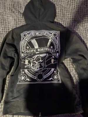 Svart hoodie med Pacific Northwest-tryck - Cool svart hoodie med dragkedja från Northern Stage Co. Stor ryggprint med texten 'Pacific Northwest' och grafiskt motiv i vitt. Utrustad med huva och två fickor framtill. Perfekt för dig som gillar streetwear och unika tryck.