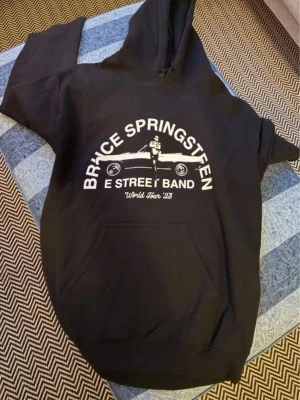 Bruce Springsteen hoodie svart S - Svart hoodie med vit Bruce Springsteen & E Street Band World Tour '23-tryck framtill. Klassisk känguruficka och huva. Tillverkad i 50% bomull och 50% polyester. Perfekt för dig som gillar bandmerch och vill ha en skön och snygg hoodie. Archive outfit med denna hoodien på den är tryckt 2023 och kunde endast fås på plats under turnén!