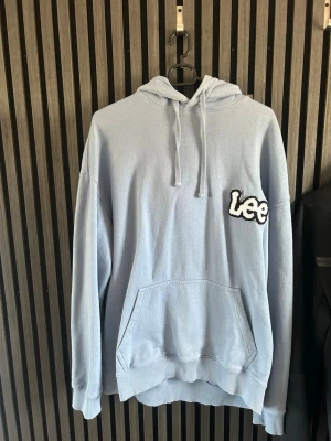 Ljusblå hoodie från Lee - Säljer en ljusblå hoodie från Lee med stor magficka och broderad Lee-logga i vitt och svart på bröstet. Hoodien har huva med dragsko och ribbade muddar vid ärmslut och nederkant. Perfekt för en chill och avslappnad stil.