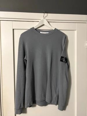 Blå sweatshirt från Calvin Klein Jeans - Snygg blå sweatshirt från Calvin Klein Jeans med rund halsringning och ribbade muddar. Tröjan har en svart CK-logga på ärmen och är tillverkad i ett mjukt, strukturerat material. Perfekt för en avslappnad och stilren look.