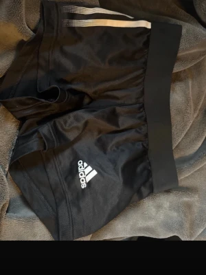 Svarta Adidas shorts med vita ränder - Säljer ett par svarta Adidas shorts med vita ränder på sidan och klassisk logga framtill. De har bred resår i midjan och är tillverkade i ett lätt och sportigt polyestermaterial. Perfekta för träning eller sportiga dagar.