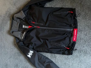 Svart vindjacka från Helly Hansen - Svart vindjacka från Helly Hansen med röda detaljer och dragkedjor. Jackan har två sidofickor med dragkedja, hög krage och logga på ärmen samt vid nederkanten. Perfekt för blåsiga dagar.