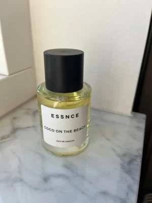 ESSNCE Coco On The Beach parfym - Fräsch Eau de Parfum från ESSNCE med namnet Coco On The Beach. Flaskan är rund och genomskinlig med svart lock och vit etikett med svart text. Innehållet är ljusgult och flaskan har en stilren, minimalistisk design som passar perfekt på parfymhyllan.