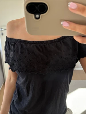 Svart offshoulder topp med spets - Säljer en svart offshoulder topp med broderad spetsvolang längs axlarna. Toppen har en luftig passform och är perfekt för varma dagar. Materialet känns mjukt och bekvämt mot huden.