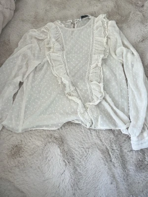 Vit volangblus från Zara - Söt vit blus från Zara med transparent tyg och diskret prickmönster. Blusen har långa ärmar och dekorativa volanger framtill samt spetsdetaljer längs kanterna. Perfekt för dig som gillar romantisk och feminin stil.