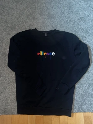 Svart sweatshirt från Ellesse - Svart sweatshirt från Ellesse med färgglatt logotryck på bröstet där bokstäverna droppar i regnbågens färger. Tröjan har rund halsringning, långa ärmar och ribbade muddar vid ärmslut och nederkant. Perfekt för en avslappnad och trendig look.