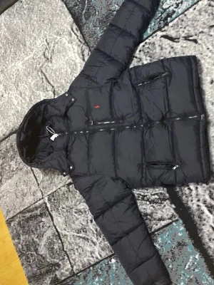 Svart pufferjacka från Polo Ralph Lauren - Svart pufferjacka från Polo Ralph Lauren med huva och flera dragkedjeförsedda fickor. Jackan har ett rött Polo-logo broderat på bröstet och är tillverkad i polyester. Perfekt för kalla dagar och har en snygg, modern look.