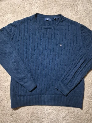 Gant stickad - Kabelstickad tröja från Gant i mörkblått med rund halsringning och diskret logga på bröstet. Tröjan har långa ärmar och ribbade muddar vid ärmslut och nederkant. Perfekt för lager på lager och klassisk stil.