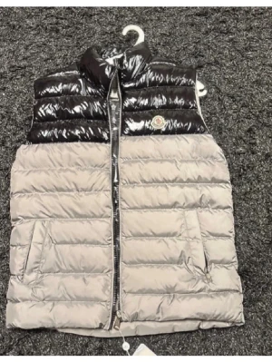 Moncler dunväst svart och grå - Snygg dunväst från Moncler i glansigt svart upptill och matt grå nedtill. Västen har hög krage, två fickor med dragkedja och klassisk Moncler-logga på bröstet. Perfekt för lager-på-lager och streetstyle.