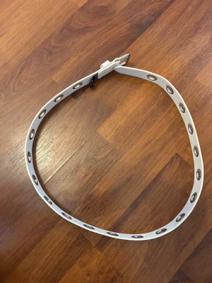 Vitt bälte med nitar från H&M Divided - Snyggt vitt bälte från H&M Divided med silverfärgade metallnitar längs hela bandet. Bältet har ett klassiskt spänne i silver och är gjort i syntetmaterial. Perfekt för att ge din outfit en edgy touch.