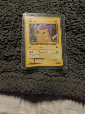 Pikachu Pokémonkort Base Set - Säljer ett klassiskt Pikachu-kort från Pokémon Base Set. Kortet har gul ram, motiv av Pikachu i en skog och är tryckt på tjockt pappmaterial. Kortet är inplastat i ett skyddsfodral och har detaljer på engelska. Perfekt för samlare eller Pokémonfans!