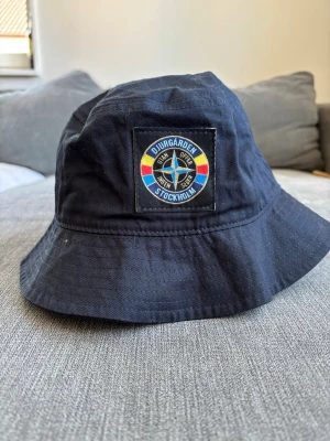 Djurgården bucket hat marinblå - Snygg marinblå bucket hat från Djurgården med broderad patch framtill i blått, gult och rött. Klassisk form med bred skärm och mjukt tyg, perfekt för dig som vill visa vilket lag du håller på. Materialet känns slitstarkt och bekvämt.