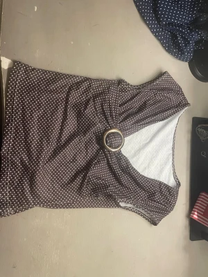 Brun prickig omlottopp med ringdetalj - Snygg brun omlottopp med vita prickar och en cool ringdetalj framtill. Toppen är ärmlös och har en figurnära passform som framhäver midjan. Perfekt för dig som gillar retrovibbar och vill sticka ut lite extra.