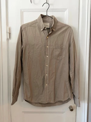 Beige skjorta från A Days March - Klassisk beige skjorta från A Days March med lång ärm, bröstficka och knappar hela vägen fram. Skjortan har button-down krage och är tillverkad i ett tunt bomullstyg som passar perfekt till lager på lager. Enkel och stilren design. Sparaamr använd. 