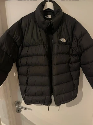 Svart pufferjacka från The North Face - Säljer en svart pufferjacka från The North Face med hög krage och dragkedja framtill. Jackan har två sidofickor med dragkedja och klassisk logga på bröstet och ryggen. Har fixat ärmen som det syns på bilden, svårt att se märkena dock! 